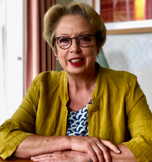 smiling woman wearing glasses and yellow jacket sitting at a table in a cozy room with natural light showcasing four elegant style elements